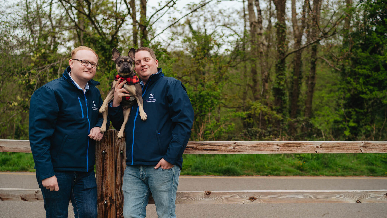 The Ruby Reese Team: Chris, Killian and Ruby the French Bulldog in a park in front of a wooden fence
