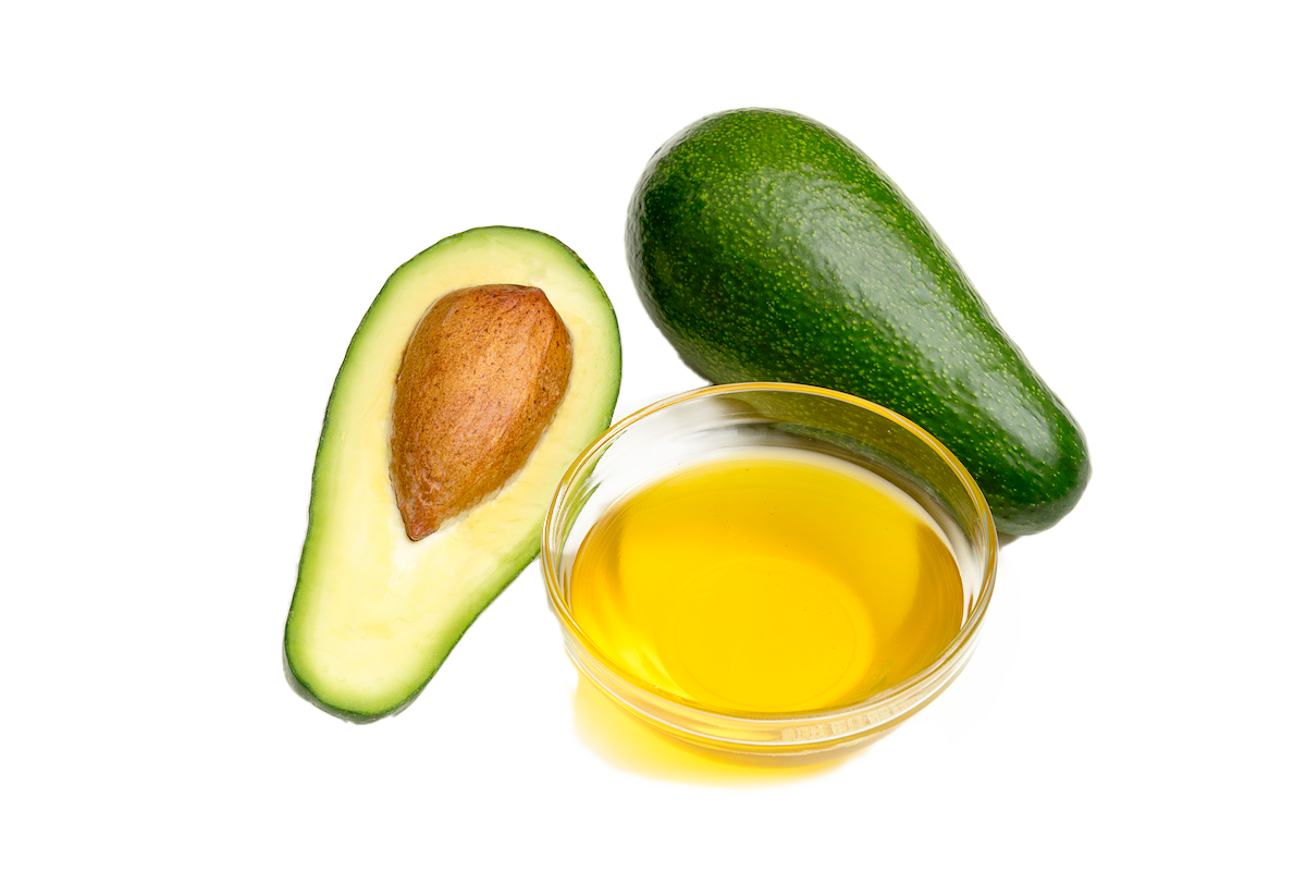 A small glass bowl filled with avocado oil. A halved avocado with the stone and a whole avocado next to it.