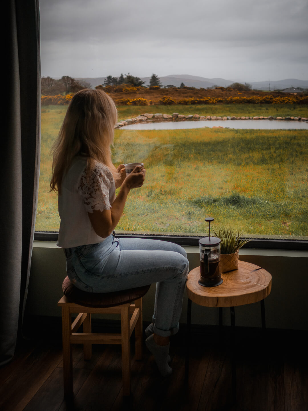 A women sitting in front of a big window watching the Irish landscape and drinking a coffee.